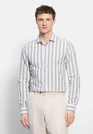 Man draagt een wit-grijs verticaal gestreept overhemd met lange mouwen, ingestopt in beige broek, staand tegen een effen lichte achtergrond.