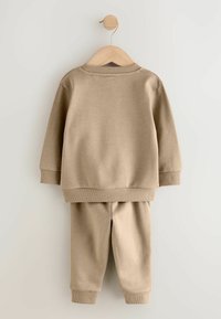 Beiger Baumwoll-Sweatshirt und passende Jogginghose mit gerippten Bündchen und Saum, präsentiert an einem Holzkleiderbügel vor einem hellen Hintergrund.