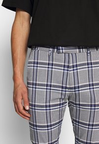 Harmaat ruudulliset housut, joissa on tummansinisiä ja valkoisia raitoja, jotka ovat sileät, vyölenkeillä ja hyvin istuvat. Yllä musta lyhythihainen paita.