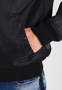 Svart, lätt bomberjacka med ribbade ärmslut, en knappficka och mjuk textur, delvis visad på en persons hand.