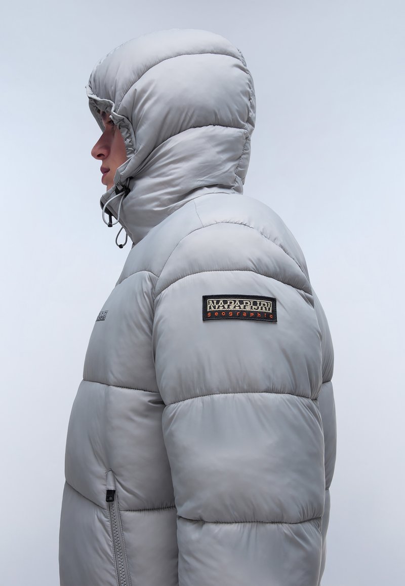 Person, der eine hellgraue gepolsterte Kapuzenjacke mit dem Napapijri Geographic-Logo auf dem Ärmel trägt und nach links vor einem einfarbigen Hintergrund blickt.