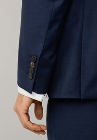Veste de costume bleu marine avec un motif texturé, présentant quatre boutons marron au poignet et les poignets de chemise blanche visibles en dessous.