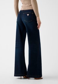 Jean large bleu foncé en denim, avec des coutures contrastantes, deux poches arrière et un patch logo sur la poche droite.