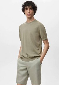 Tricou cu mânecă scurtă și guler rotund, de culoare verde măsliniu deschis, fabricat dintr-un material tricotat, purtat cu pantaloni scurți asortați de culoare deschisă. Textură netedă, croială lejeră.