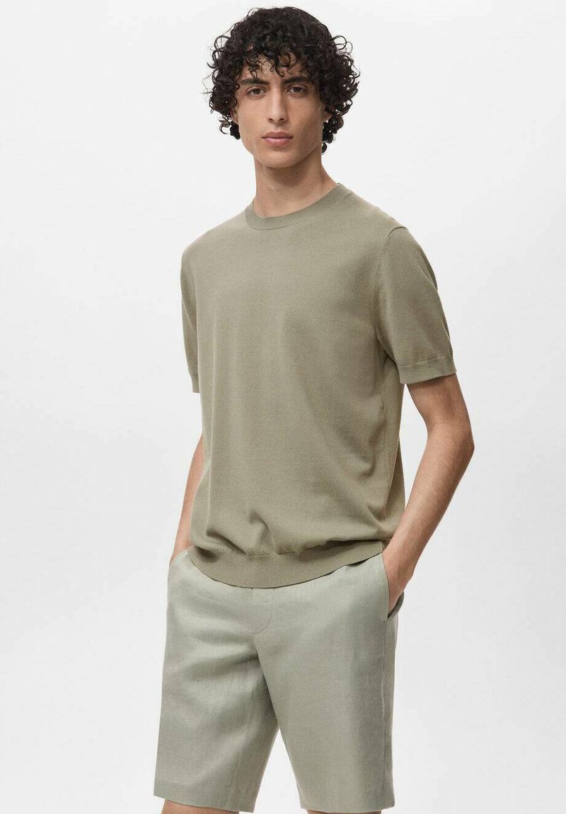 Tricou cu mânecă scurtă și guler rotund, de culoare verde măsliniu deschis, fabricat dintr-un material tricotat, purtat cu pantaloni scurți asortați de culoare deschisă. Textură netedă, croială lejeră.