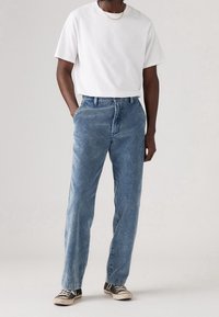 Homme portant un t-shirt blanc, un pantalon en velours côtelé bleu clair, des baskets noires et blanches, debout les mains dans les poches sur un fond uni.