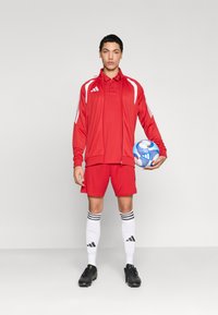 Κόκκινη αθλητική εμφάνιση που περιλαμβάνει ζακέτα με φερμουάρ, πόλο μπλούζα και σορτς. Μαύρα ποδοσφαιρικά παπούτσια και λευκές κάλτσες μέχρι το γόνατο με μαύρες ρίγες. Κρατάει μια μπάλα ποδοσφαίρου.