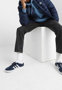 Marineblå puffjacka över en blå hoodie, matchad med mörka jeans och marinblå sneakers med vita ränder. Modell sitter på en vit kub.