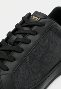 Schwarzer Ledersneaker mit glatter Oberfläche und strukturiertem Muster, schwarzen Schnürsenkeln und einem goldfarbenen Logoakzent auf der Zunge.