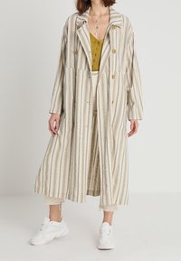 Pruhovaný dlouhý kabát v béžové, modré a krémové barvě. Má vykrojený límec, dvouřadé zapínání a velké knoflíky. Nosený přes hořčicový top.