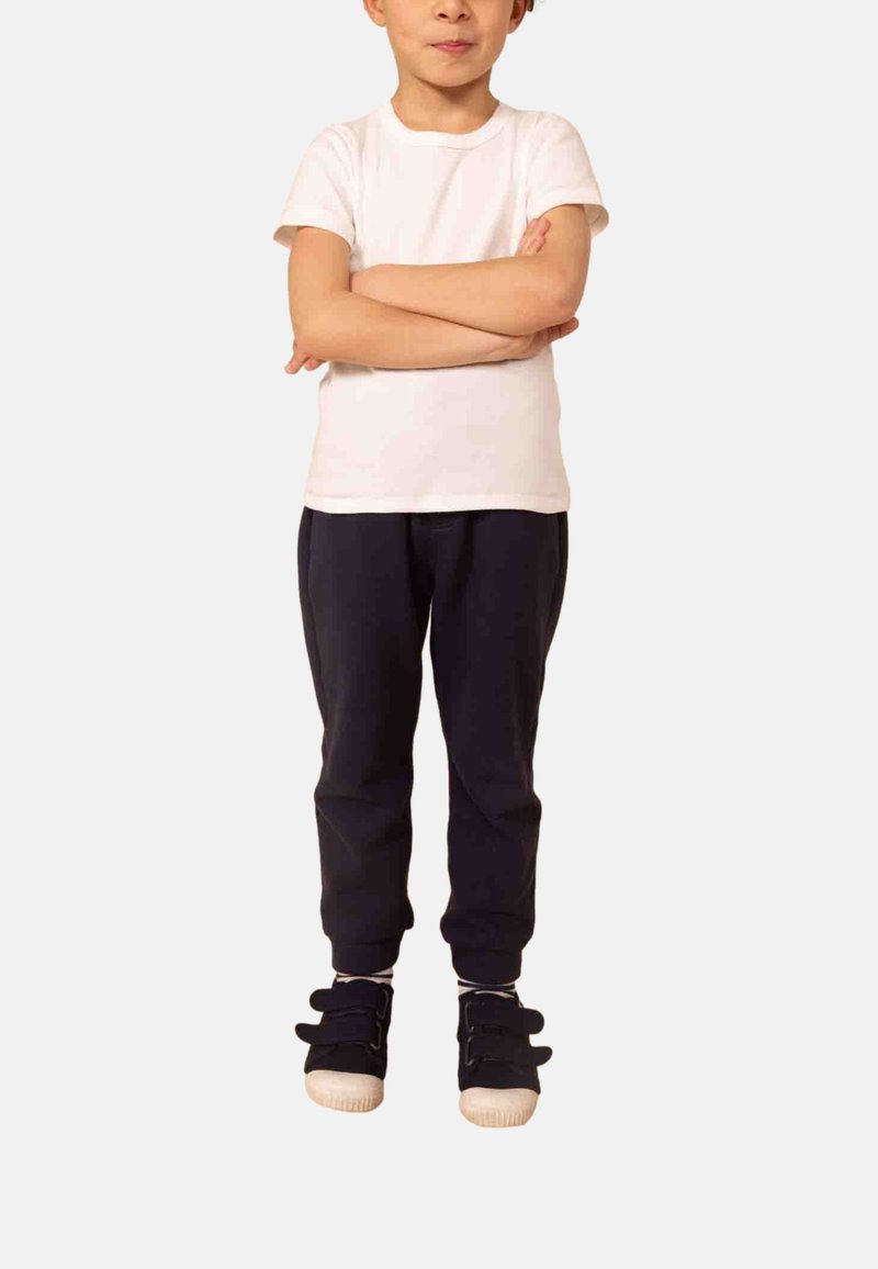 Barn iført en hvid kortærmet skjorte, sorte bukser og sorte velcrosko, stående med armene over kors mod en ensfarvet baggrund.