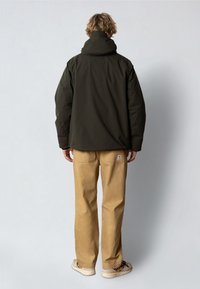 Tummanvihreä takki, jossa on korkea kaulus, pehmeä kangas ja rento leikkaus, yhdistettynä vaaleanbeigeihin housuihin ja rentoihin jalkineisiin.