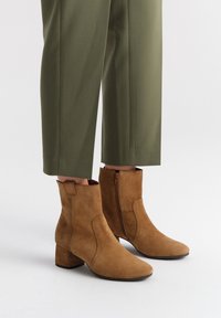 Brązowe zamszowe botki za kostkę na słupku, z bocznym zamkiem błyskawicznym, spiczastym noskiem i gładką teksturą, w połączeniu z oliwkowymi spodniami.