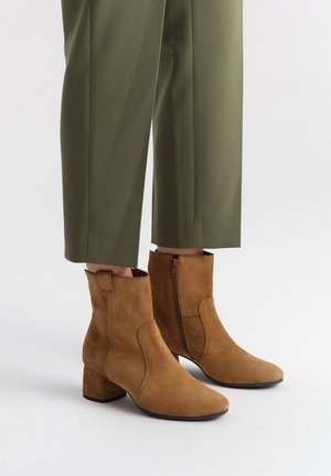 Brązowe zamszowe botki za kostkę na słupku, z bocznym zamkiem błyskawicznym, spiczastym noskiem i gładką teksturą, w połączeniu z oliwkowymi spodniami.