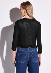 Musta, puoliksi läpikuultava neulepaita, jossa on kolme neljästä hihasta, yhdistettynä sinisiin, korkeavyötäröisiin farkkuihin, joissa on takataskut ja vaalea ommel.