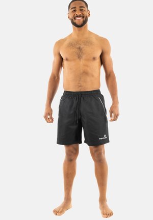 Hombre sonriente sin camiseta, de pie descalzo, vistiendo pantalones cortos negros con cordón y bolsillos, sobre un fondo liso claro.