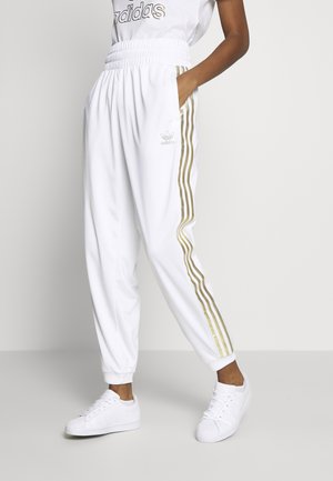 Personne portant un pantalon de survêtement Adidas blanc avec des bandes latérales dorées, des baskets blanches et un t-shirt Adidas blanc, debout avec une main dans la poche.