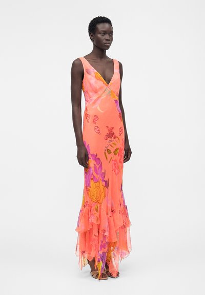 Γυναίκα που φοράει φορεμα χωρίς μανίκια μέχρι το πάτωμα σε κοραλλί χρώμα με ροζ, πορτοκαλί και μοβ λουλουδένια σχέδια και βολάν στη φούστα, στέκεται μπροστά σε απλό φόντο.