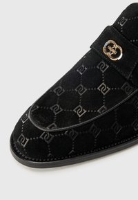 Mocassins en suède noir présentant un motif texturé, une garniture en cuir, et un détail en métal doré sur la sangle. Semelle basse.