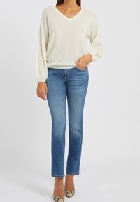Pull en ivoire à col en V avec des manches longues, associé à un jean bleu droit. Le mannequin porte des escarpins à imprimé serpent, mettant en avant des textures douces et un style décontracté.