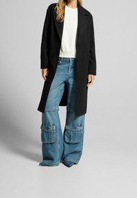 Cappotto nero a righe, felpa bianca, pantaloni in denim blu a gamba larga con tasche cargo e scarpe blu a punta. Tessuto strutturato e look stratificato.