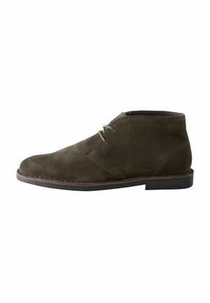 Botín chukka de ante verde oliva con puntera redondeada, cordones simples y suela de goma negra. Presenta detalles cosidos a lo largo del empeine.