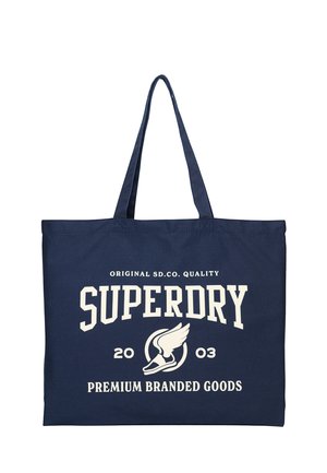 Bolsa de mano azul marino con asas largas, con texto blanco "SUPERDRY" y un logo de zapato alado con "2003" y "Premium Branded Goods" impresos en la parte frontal.