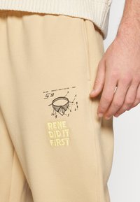Beige sweatpants van een gestructureerde stof hebben een handgetekende illustratie en gele geborduurde tekst "RENE DID IT FIRST."