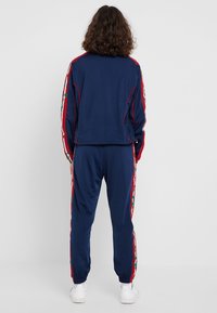 Person står vänd bort med en marinblå träningsdräkt med röda och blommiga sidstreck, i kombination med vita sneakers, mot en enkel vit bakgrund.