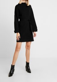 Svart ribbad klänning i tröja med rund halsringning, långa ärmar och knytmidja. Matchas med svarta ankelstövlar med en elegant finish.