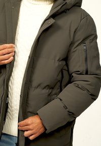 Grüne, gepolsterte Jacke mit Kapuze, die über eine Reißverschlusstasche am Ärmel und einen Reißverschluss vorne verfügt, getragen über einem cremefarbenen, grob gestrickten Pullover.