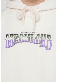 Krémová mikina s kapucí a šňůrkami, s grafickým potiskem "DREAMLAND" v černé a fialové barvě a textem "KAOTIKO SOCIETY" nad ním v černé barvě.