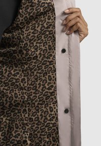 Veste de couleur claire avec une doublure en imprimé léopard. La veste présente une texture lisse et comporte des boutons pression noirs visibles.