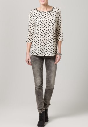 Kobieta ubrana w biały top z czarnym wzorem serc, szare przetarte wąskie jeansy, czarne botki oraz srebrną bransoletkę na prawej ręce.