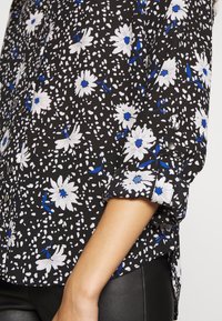 Černá květinová košile s bílými sedmikráskami a modrými akcenty, volného střihu, s knoflíkovými detaily na rukávech a jemnou, texturovanou tkaninou.