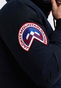 Veste marine avec un patch rond rouge et bleu bien visible sur la manche, portant l'inscription « JACKIT MOUNTAIN RANGE » et des détails de design géométrique.