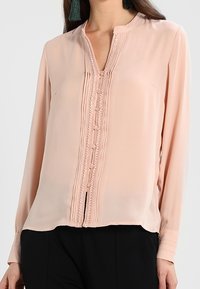 Blusa rosa chiaro con scollatura rotonda, dettagli con bottoni e pieghe. Il modello la indossa con pantaloni scuri, mettendo in evidenza uno stile casual ed elegante.