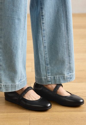 Schwarze Leder-Ballettschuhe mit runder Zehe und flexibler elastischer Riemen, kombiniert mit hellblauen, weit geschnittenen Jeans auf einem Holzboden.