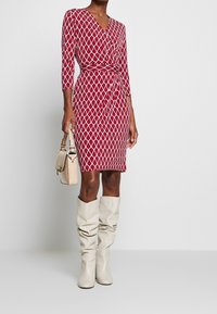 Robe portefeuille rouge avec motif géométrique blanc, manches trois-quarts, taille nouée, associée à des bottes beige souples montant jusqu'aux genoux et un petit sac.