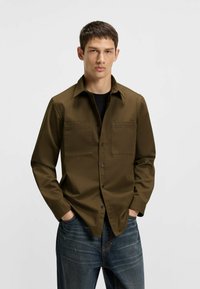 Olive green button-up shirt in a smooth fabric with two chest pockets, worn over a black shirt and paired with dark blue jeans.