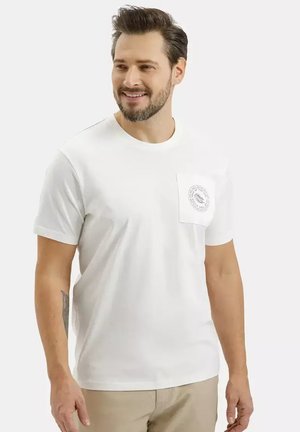 Uśmiechnięty mężczyzna w białym t-shircie z okrągłym logo na lewej kieszeni na piersi i beżowych spodniach, stojący na jednolitym tle.