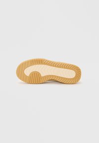 Semelle d'une chaussure, présentant une surface en caoutchouc texturée, de couleur brun clair, avec des motifs de zigzag et une section centrale lisse.