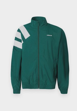 Zaļš Adidas vējjaka ar baltu logotipu uz krūtīm, baltām diagonālām svītrām uz kreisā pleca, priekšējo rāvējslēdzēju un elastīgām aprocēm un apakšu.