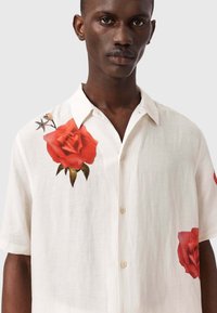 Hombre que lleva una camisa blanca de manga corta con botones y grandes estampados de rosas rojas en el pecho y en la parte inferior lateral.