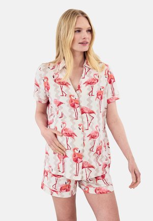 Kobieta ubrana w pasującą koszulkę z krótkim rękawem i szorty z nadrukiem różowych flamingów na jasnym tle, stojąca z jedną ręką na biodrze.