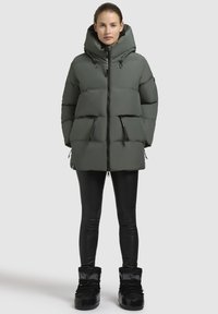 Gevoerde, olijfgroene parka met een grote capuchon, twee voorzakken met rits en een zwarte ritssluiting. Gemaakt over zwarte leggings en laarzen.