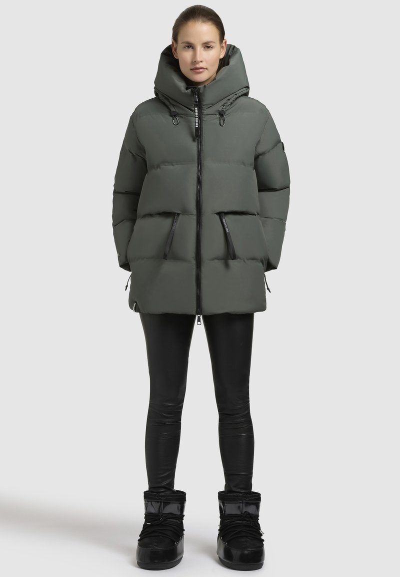 Gevoerde, olijfgroene parka met een grote capuchon, twee voorzakken met rits en een zwarte ritssluiting. Gemaakt over zwarte leggings en laarzen.