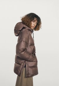 Kvinde med krøllet hår iført en brun polstret hættetrøje og beige nederdel, der kigger over skulderen mod en ensfarvet baggrund.