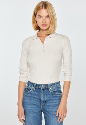 Blondynka w kremowej, długorękawowej, prążkowanej koszulce polo i niebieskich, wysokich dżinsach stoi na jednolitym jasnym tle.
