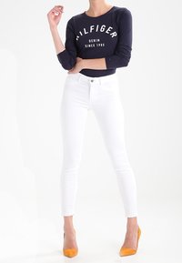 Námořnická dlouhá košile s bílým textem, v kombinaci s bílými skinny džíny a žlutými vysokými podpatky. Zaměřeno na látku a přiléhavý design.